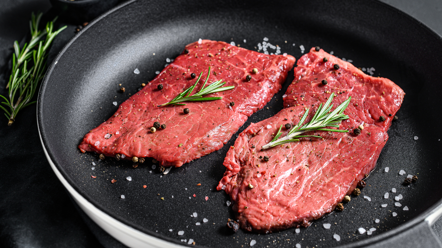 Raw seasoned steaks in a black skillet with fresh rosemary and cracked black pepper.