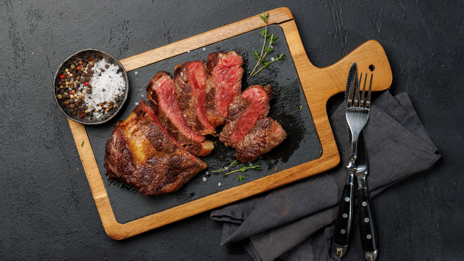Sliced ribeye steak served on a wooden board with fresh herbs and seasoning.