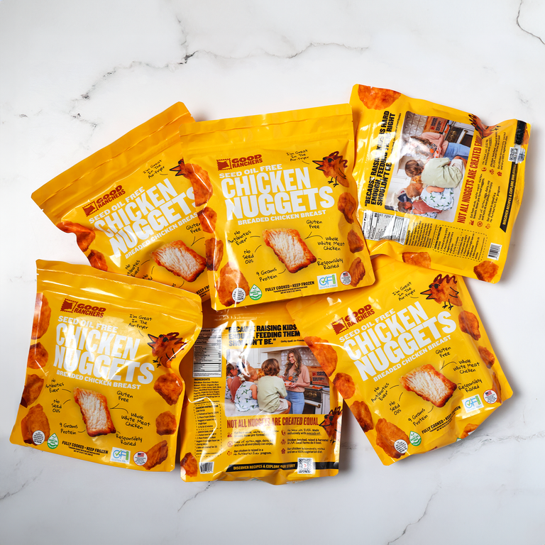 Five yellow bags of Seed Oil Free Chicken Nugget Bundle, now gluten-free and seed oil free, are displayed on a white marble surface. The packaging shows nuggets, kids eating, and product details.