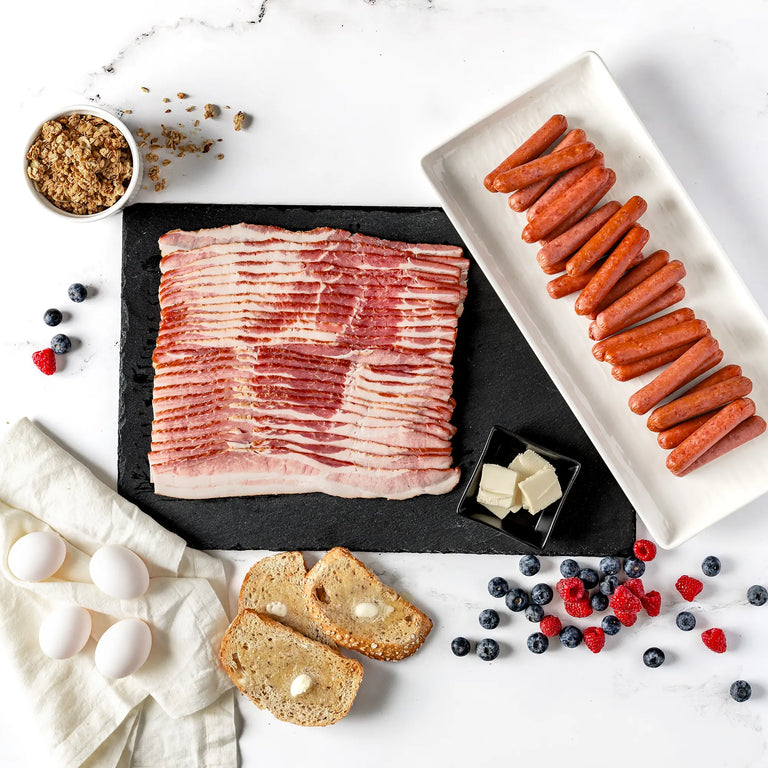 A flat lay of the Big Breakfast Bundle: Applewood bacon, breakfast sausage, cubed butter, three eggs, buttered bread slices, granola, blueberries, and raspberries on a white background.