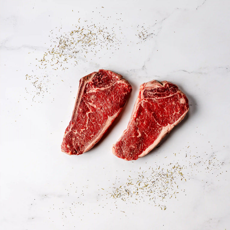 Two raw ribeye steaks on a white surface with seasoning.