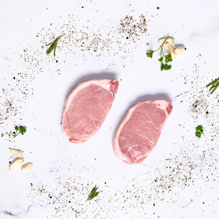 Boneless Pork Chops on a white surface, surrounded by black pepper, fresh parsley, garlic cloves, and rosemary sprigs.