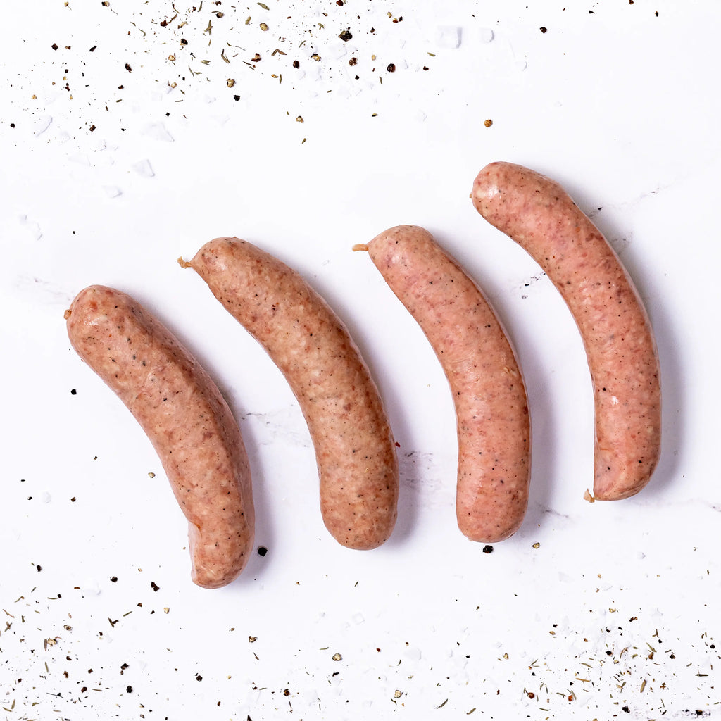 Four uncooked Smoked Brats are arranged diagonally on a white surface, surrounded by scattered grains of salt and cracked black pepper.