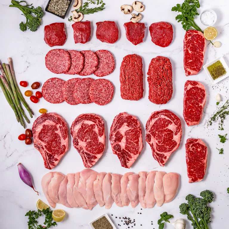 An overhead view of the Good Ranchers Butchers Bundle—free range chicken breasts, steaks, ground beef, and American wagyu burger patties—arranged neatly on a white surface with fresh herbs, vegetables, and spices.