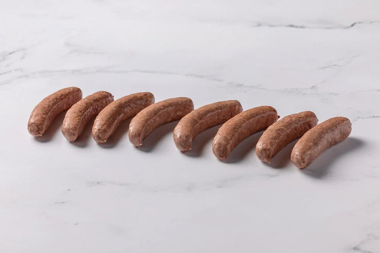 Four uncooked Smoked Brats are arranged diagonally on a white surface, surrounded by scattered grains of salt and cracked black pepper.