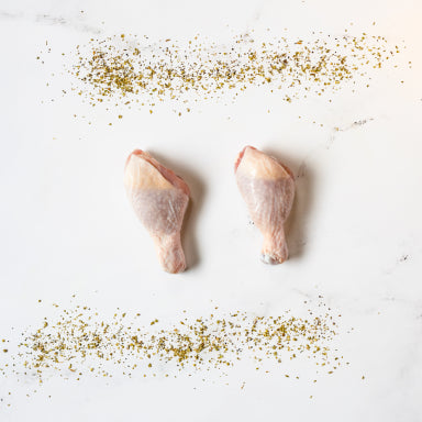Two Plain Chicken Drumsticks are placed side by side on a white surface, with lines of green herbs or spices sprinkled above and below them.