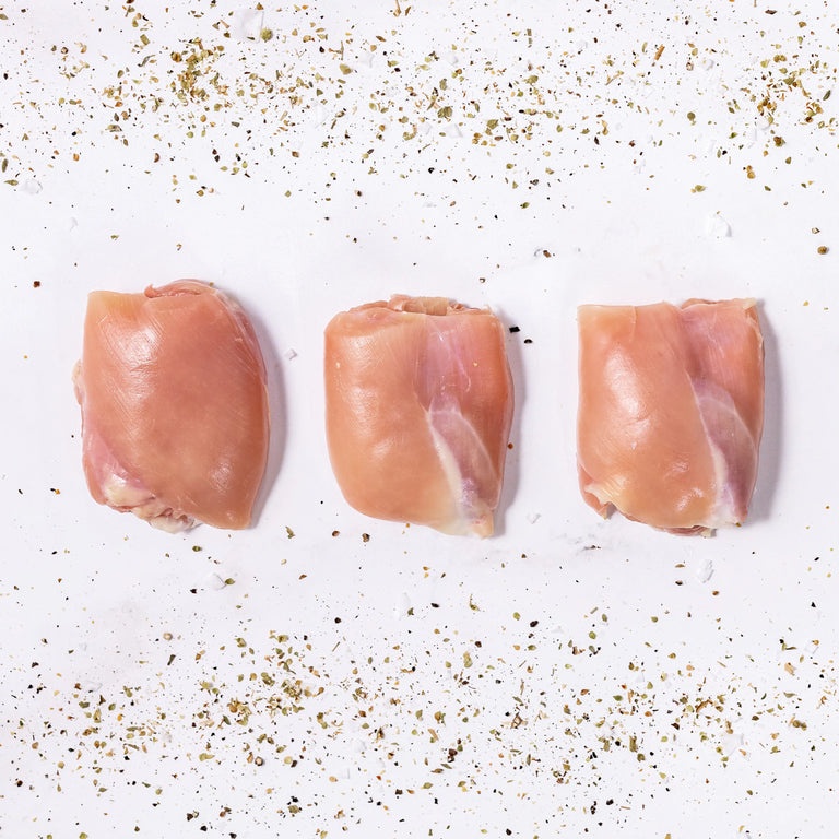 Three Boneless Skinless Chicken Thighs are arranged in a row on a white surface, surrounded by scattered dried herbs and spices.