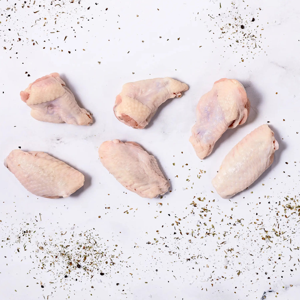 Six pieces of Chicken Wings are arranged on a white surface, with dried herbs and spices scattered around them.