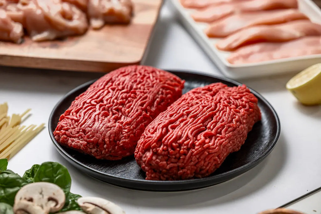 The Essentials Box by Good Ranchers has 100% American beef portions. Here, you can see it on a black plate, surrounded by spinach, mushrooms, uncooked pasta, Free Range chicken breast with no antibiotics ever, and half a lemon all in the background.