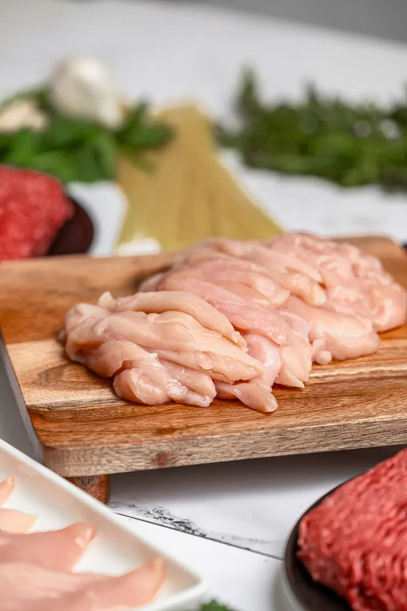 The Essentials Box features 100% American, free-range chicken breast on a wooden cutting board, with uncooked spaghetti, ground meat, herbs, and garlic in the background. This easy meal prep box contains no added hormones and delivers straight to your door.