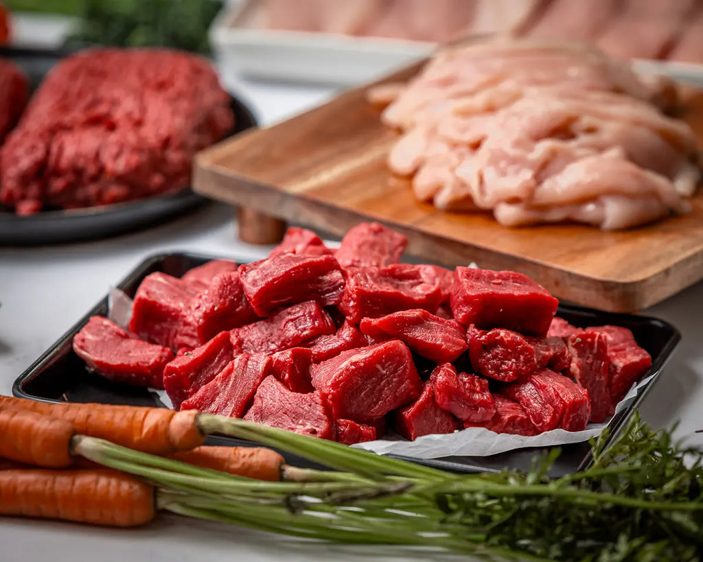 A close-up of 100% American steak tips on a tray, with carrots and other meats like 100% American ground beef and sliced chicken nearby—The Essentials Box brings these fresh cuts straight to your door.