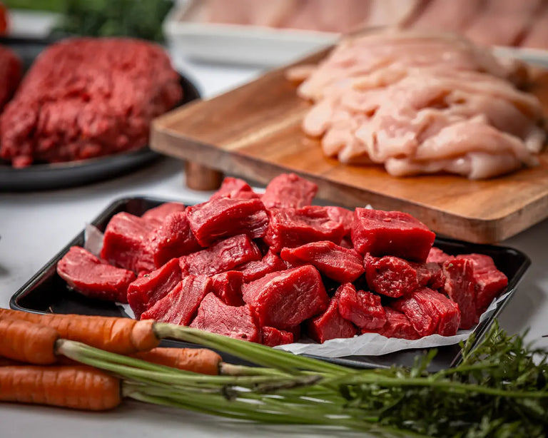 A close-up of 100% American steak tips on a tray, with carrots and other meats like 100% American ground beef and sliced chicken nearby—The Essentials Box brings these fresh cuts straight to your door.