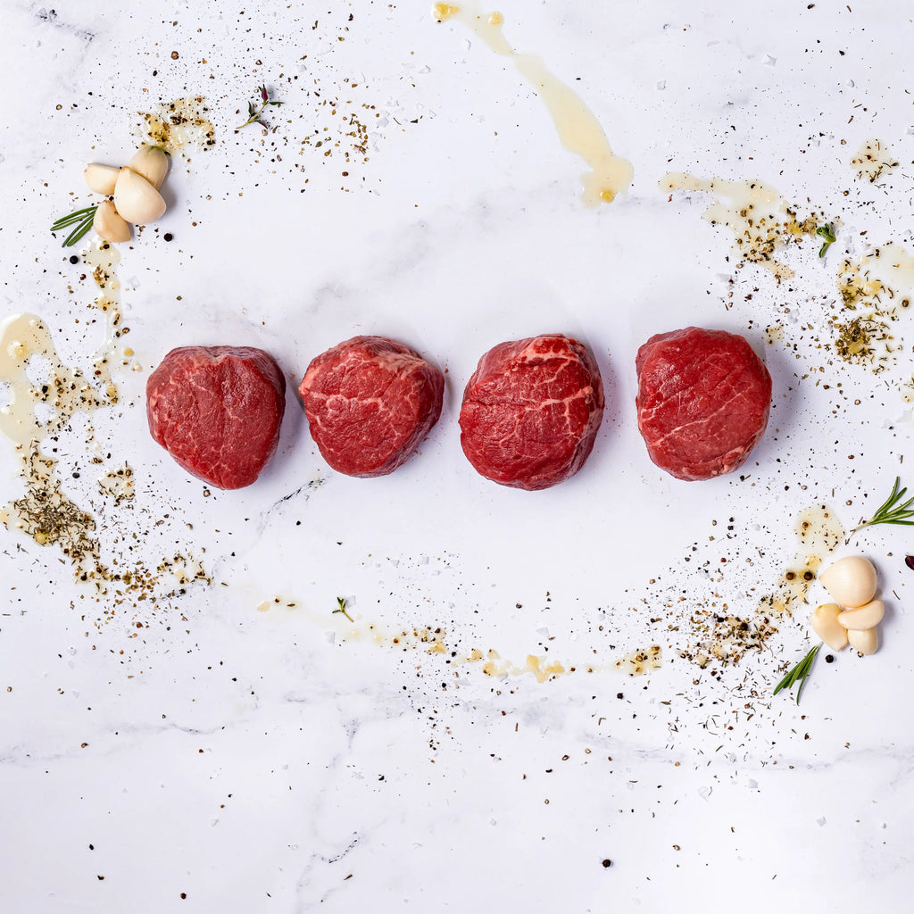 Four raw Center-Cut Filet Mignons, aged 21 days, are lined up on white marble and surrounded by scattered herbs, spices, garlic cloves, rosemary sprigs, and a drizzle of oil.