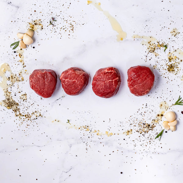 Four raw Center-Cut Filet Mignons, aged 21 days, are lined up on white marble and surrounded by scattered herbs, spices, garlic cloves, rosemary sprigs, and a drizzle of oil.