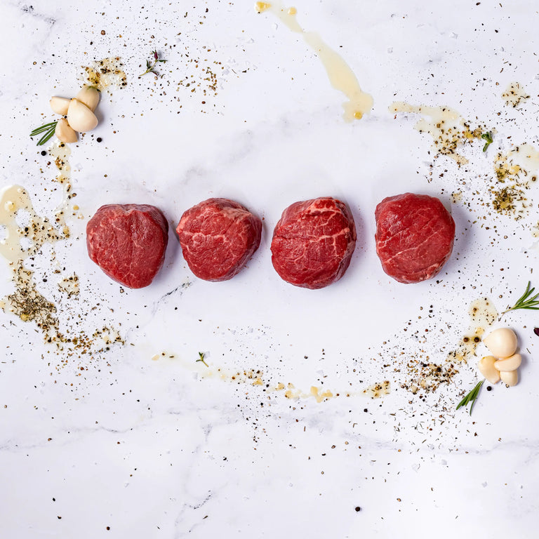 Four raw Center-Cut Filet Mignons, aged 21 days, are lined up on white marble and surrounded by scattered herbs, spices, garlic cloves, rosemary sprigs, and a drizzle of oil.