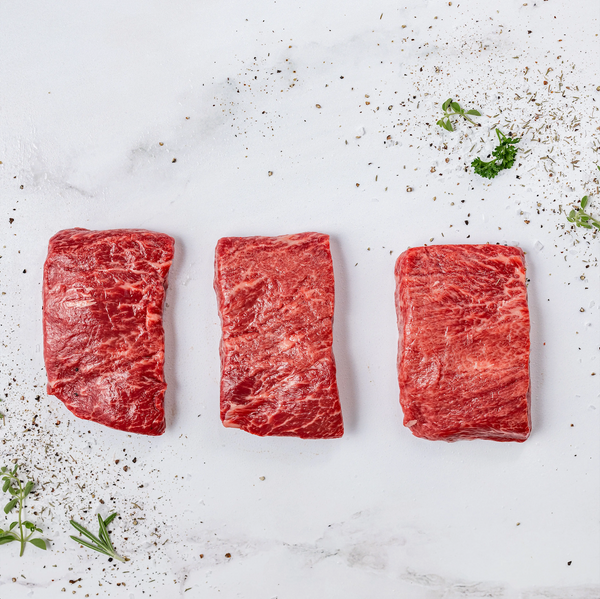 Three raw Flat Iron Steaks are arranged in a row on a white surface, surrounded by scattered pepper, herbs, and seasoning.