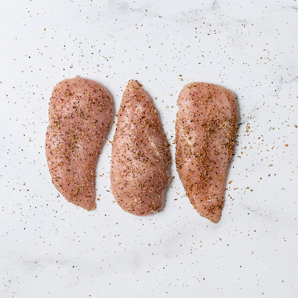Three Garlic Pepper Chicken Breasts are arranged in a row on a white marble surface, with extra seasoning sprinkled around them.