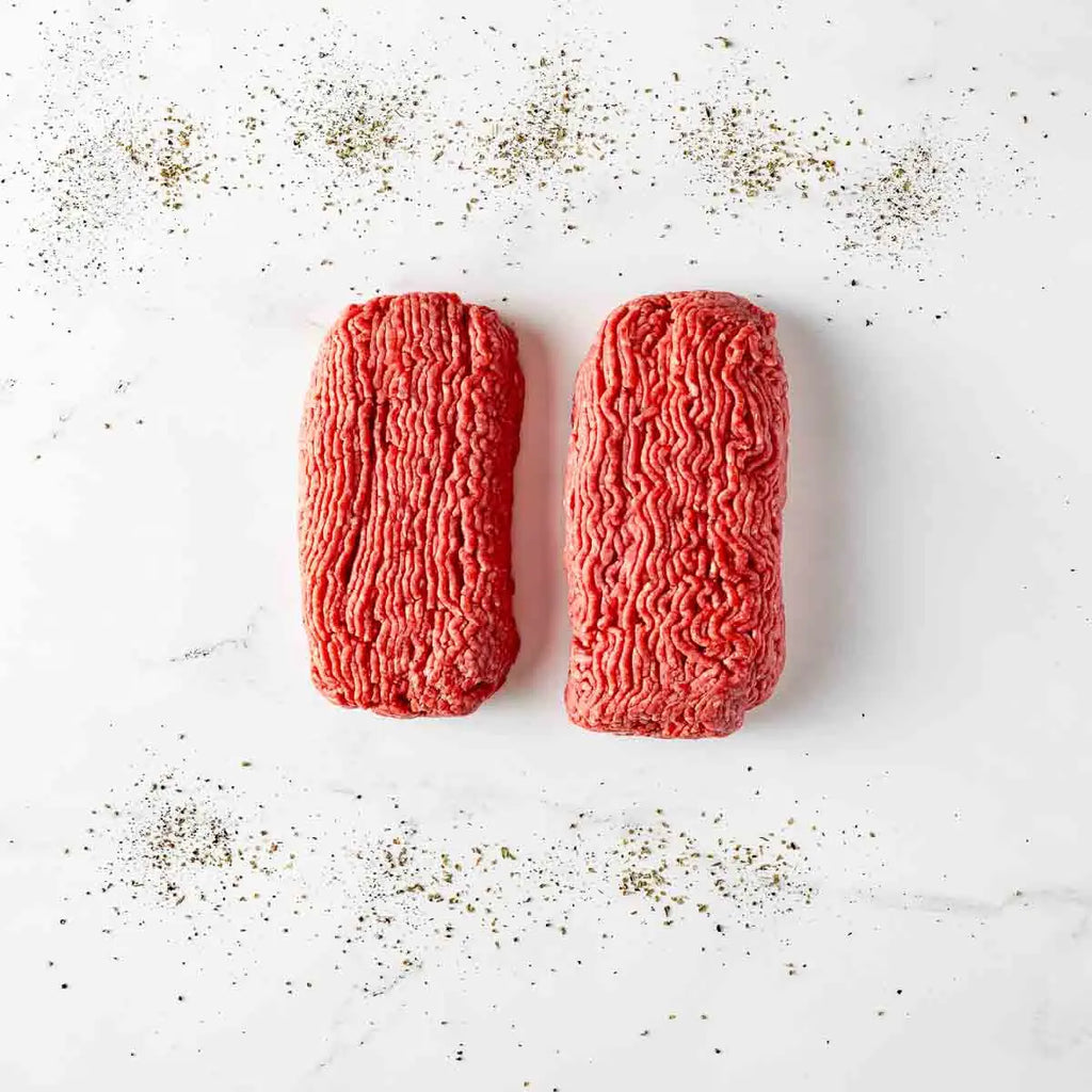 Two portions of Grass Fed Ground Beef Gift are placed side by side on a white surface, with scattered black pepper sprinkled around them.