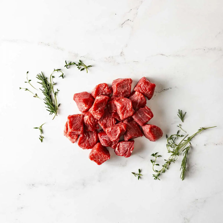 Grass Fed Beef Steak Tips Gift: Cubes of raw beef arranged on a white marble surface, garnished with sprigs of fresh rosemary and thyme.