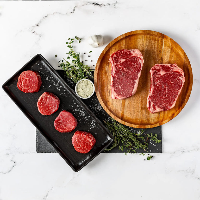 The His & Hers Steak Night Bundle features two raw ribeye steaks on a round wooden plate and five filet mignon medallions on a sleek black platter, garnished with herbs and seasoning—perfect for an elevated date night.
