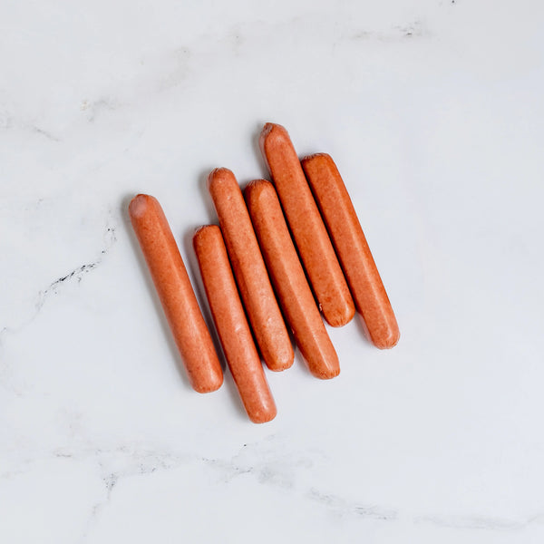 Six uncooked Hot Dogs are arranged in a slightly scattered row on a white marble surface.