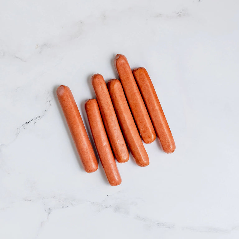 Six uncooked Hot Dogs are arranged in a slightly scattered row on a white marble surface.