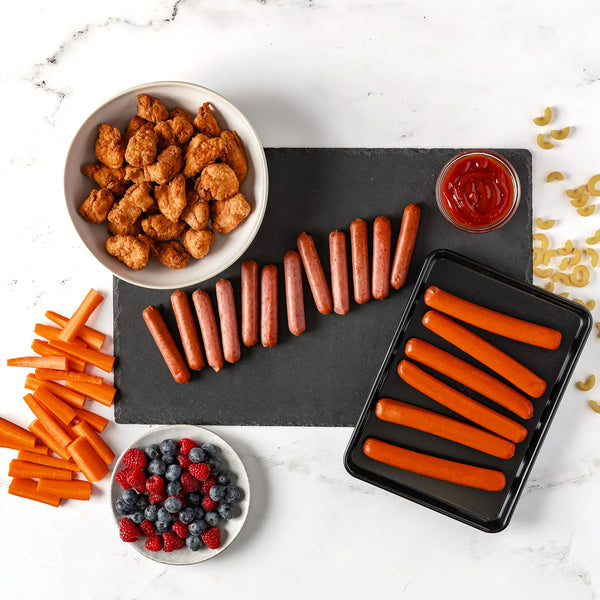 A flat lay featuring the Hungry Kids Bundle: Chicken Nuggets in a bowl, Breakfast Sausage Links on a board, Hot Dogs on a tray, plus carrot sticks, mixed berries, uncooked pasta, and ketchup on a white surface.