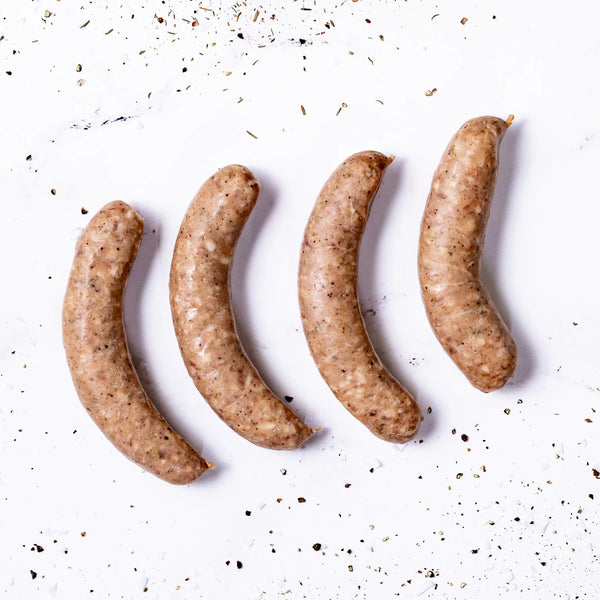 Four uncooked Italian Sausages are lined up on a white surface, sprinkled with black pepper and spices.