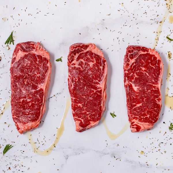 Three raw, marbled New York Strips are lined up on a white surface, topped with herbs and olive oil, and garnished with small rosemary sprigs.