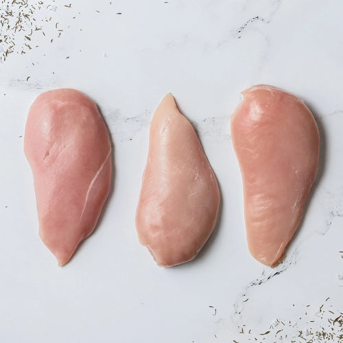 Three raw, skinless FREE Plain Chicken Breast fillets are placed side by side on a white marble surface, with herbs scattered in the corners.