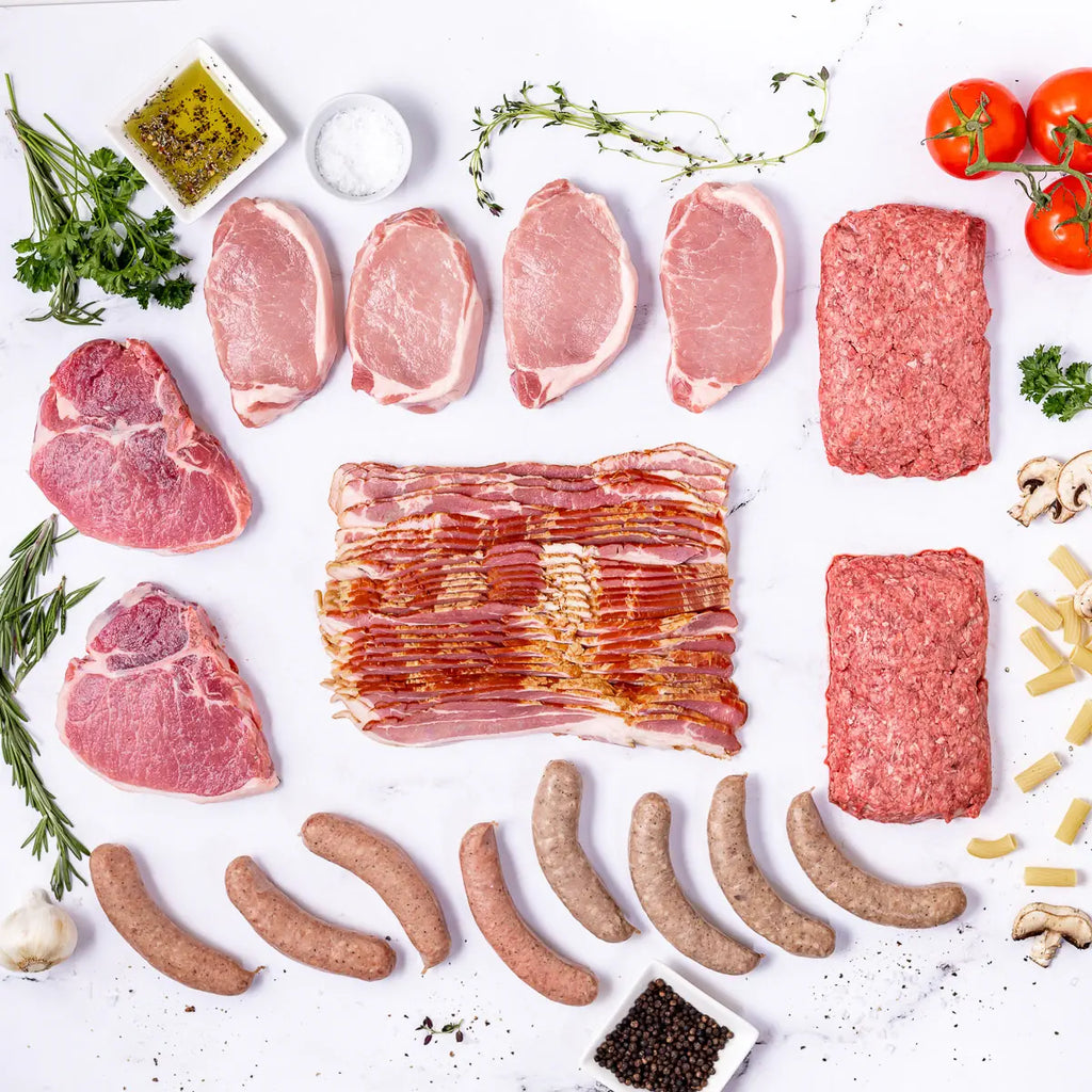 The Prime Pork Box showcases 100% American, seed oil free raw meats like bacon, pork chops, and sausages on a white surface with fresh herbs, tomatoes, mushrooms, pasta, garlic, salt, pepper around it.