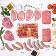 The Prime Pork Box showcases 100% American, seed oil free raw meats like bacon, pork chops, and sausages on a white surface with fresh herbs, tomatoes, mushrooms, pasta, garlic, salt, pepper around it.
