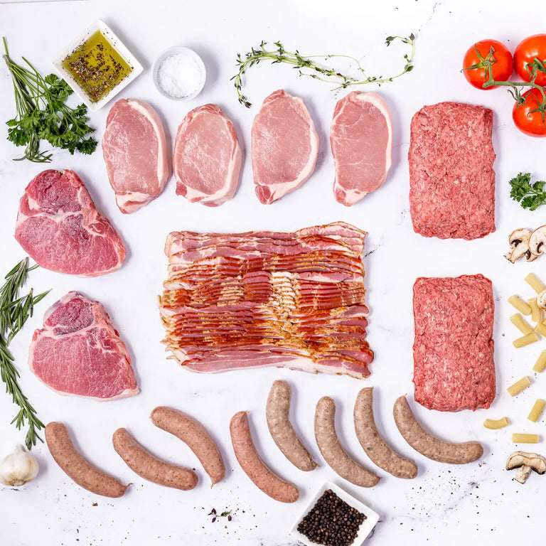 The Prime Pork Box showcases 100% American, seed oil free raw meats like bacon, pork chops, and sausages on a white surface with fresh herbs, tomatoes, mushrooms, pasta, garlic, salt, pepper around it.