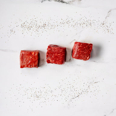 Three raw Ranch Steaks are lined up on a white marble surface and lightly sprinkled with black pepper.