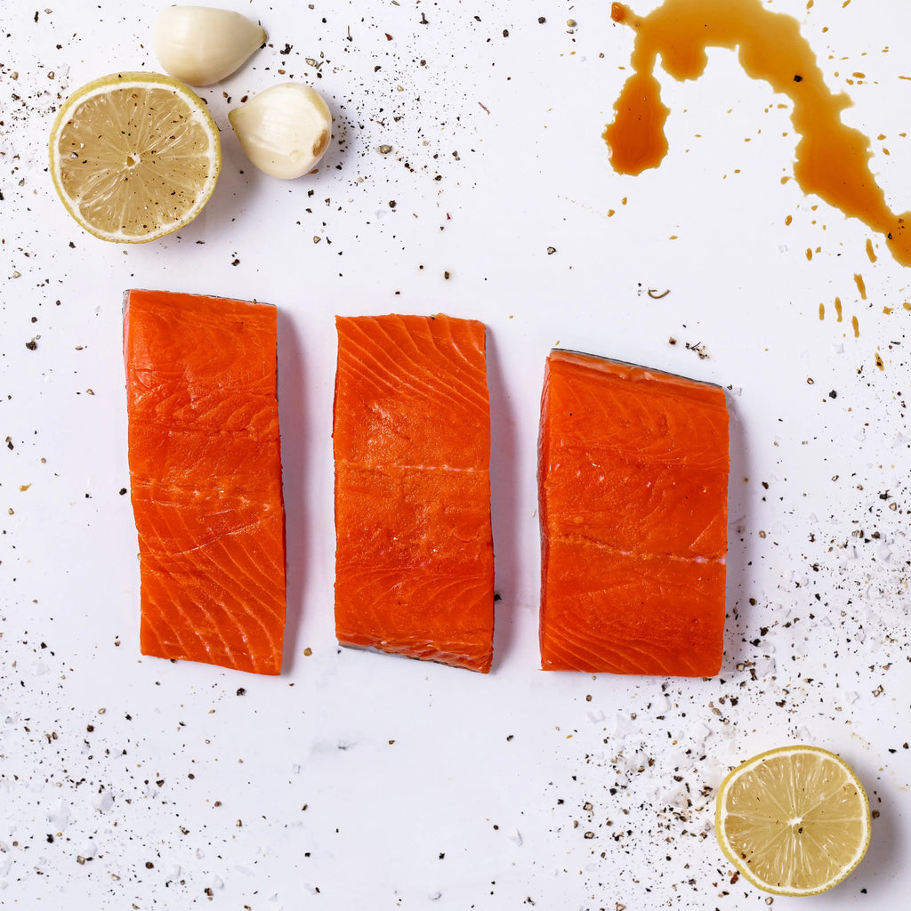 Three FREE Wild Caught Salmon fillets are arranged on a white surface with two lemon halves, two garlic cloves, scattered pepper, and a splash of sauce or marinade.