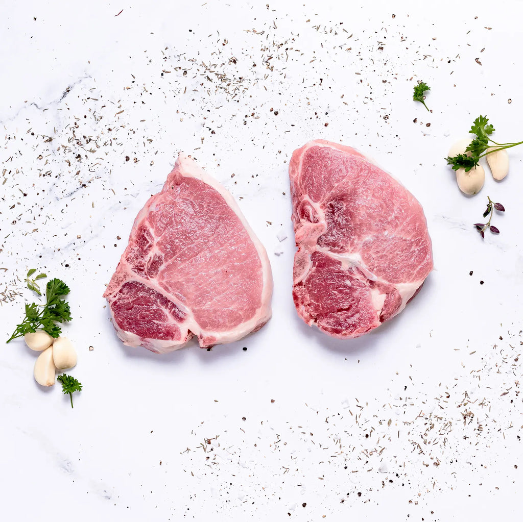 Two Steakhouse Cut Pork Chops on a white surface, surrounded by scattered herbs, parsley sprigs, and garlic cloves.