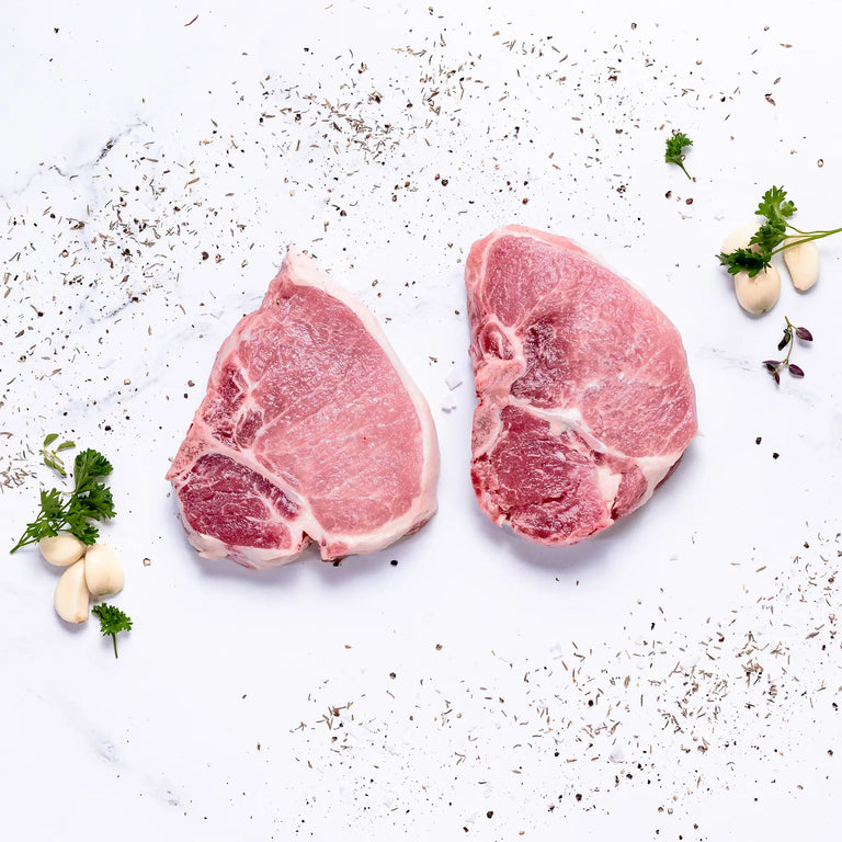 Two Steakhouse Cut Pork Chops on a white surface, surrounded by scattered herbs, parsley sprigs, and garlic cloves.