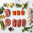 The Surf And Turf Box—featuring locally sourced steak cuts, wild-caught salmon fillets, and gulf shrimp—is artfully arranged with lemon halves, asparagus, fresh herbs, garlic, pepper, and seasoning bowls on a white surface.