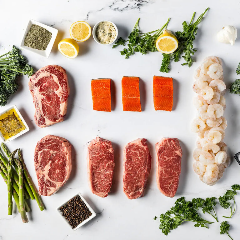 The Surf And Turf Box—featuring locally sourced steak cuts, wild-caught salmon fillets, and gulf shrimp—is artfully arranged with lemon halves, asparagus, fresh herbs, garlic, pepper, and seasoning bowls on a white surface.