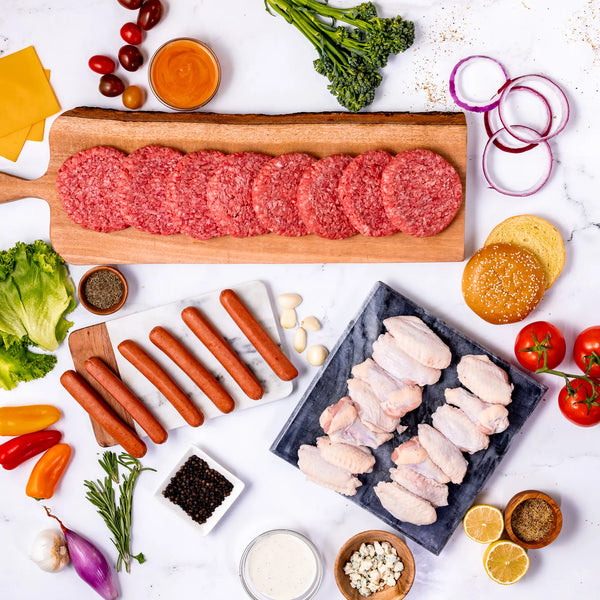 The Summer Grilling Bundle is arranged on platters with fresh veggies, cheese, buns, sauces, and seasonings on a white marble surface—perfect for your next cookout or BBQ.