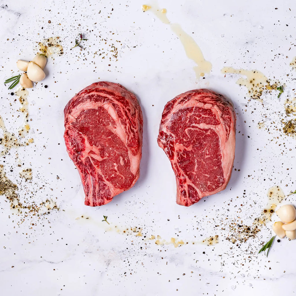 Two raw Thick-Cut, Bone-In Ribeyes, each 22oz, rest on a white marble surface surrounded by scattered herbs, black pepper, olive oil drizzles, garlic cloves, and sprigs of rosemary.