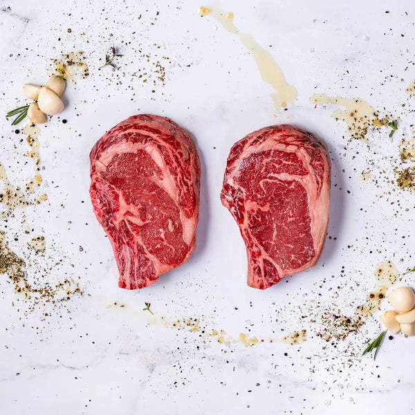Two raw Thick-Cut, Bone-In Ribeyes, each 22oz, rest on a white marble surface surrounded by scattered herbs, black pepper, olive oil drizzles, garlic cloves, and sprigs of rosemary.